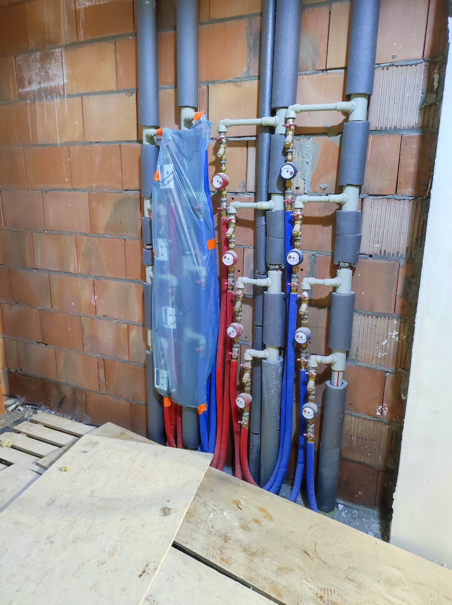 Rozdzielacz ogrzewania podłogowego w folii ochronnej, rury doprowadzające ciepło w kolorze czerwonym i powrotne w kolorze niebieskim, zaizolowane piony, widoczna surowa ściana z cegły.