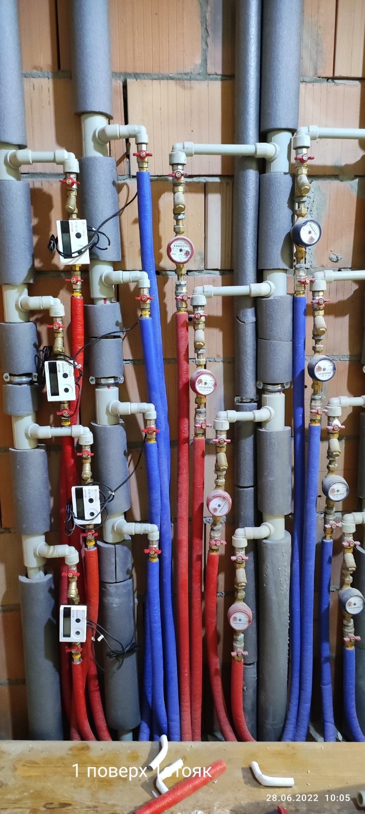 Ściana z cegły z zainstalowanymi rzędami rur grzewczych w izolacji, z zaworami i licznikami ciepła, widoczne fragmenty rur i odpadów na podłodze.