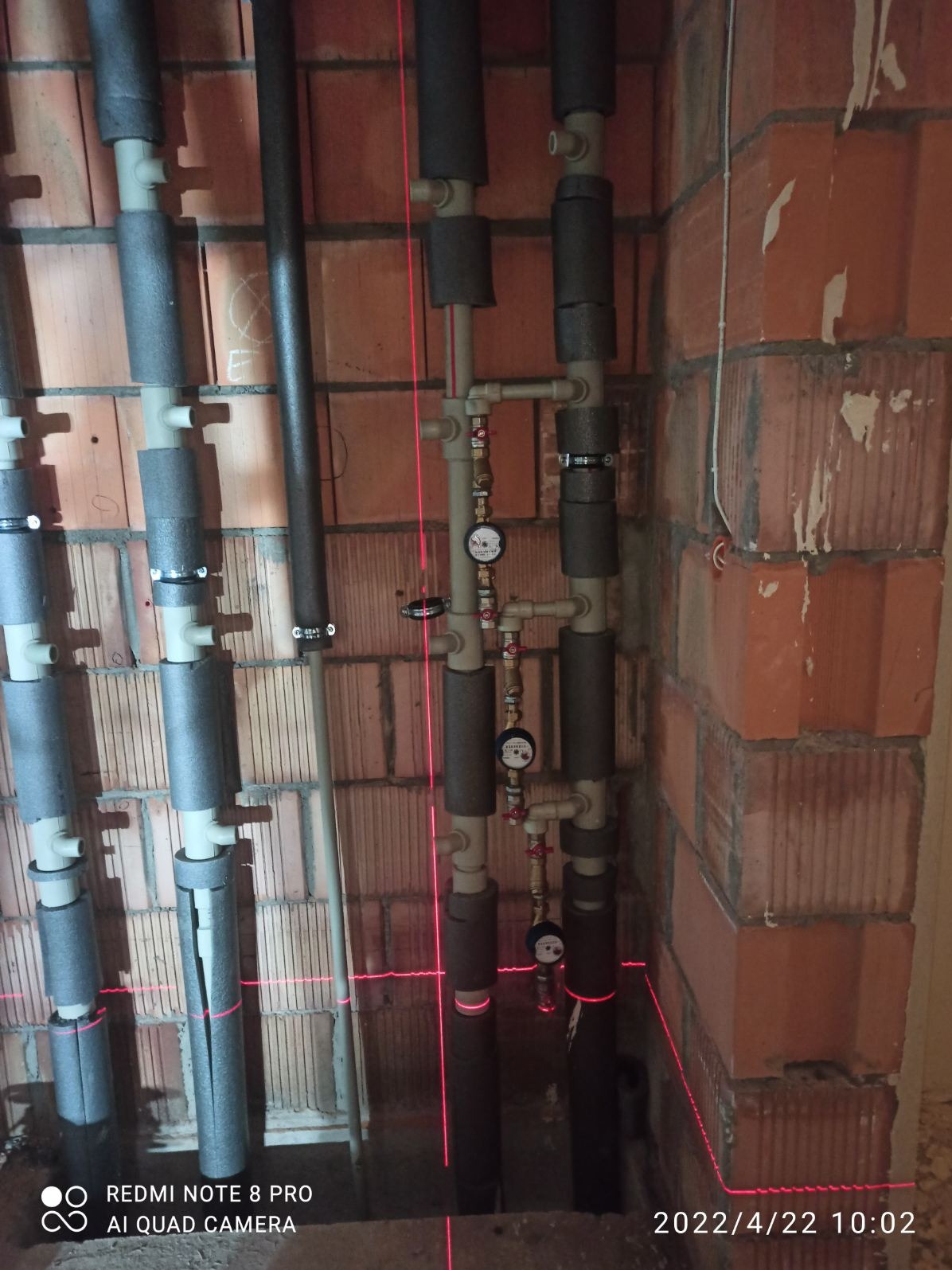 Instalacja rur grzewczych z izolacją, manometry i zawory zamontowane na ścianie z cegły, wyznaczona poziomica laserowa.