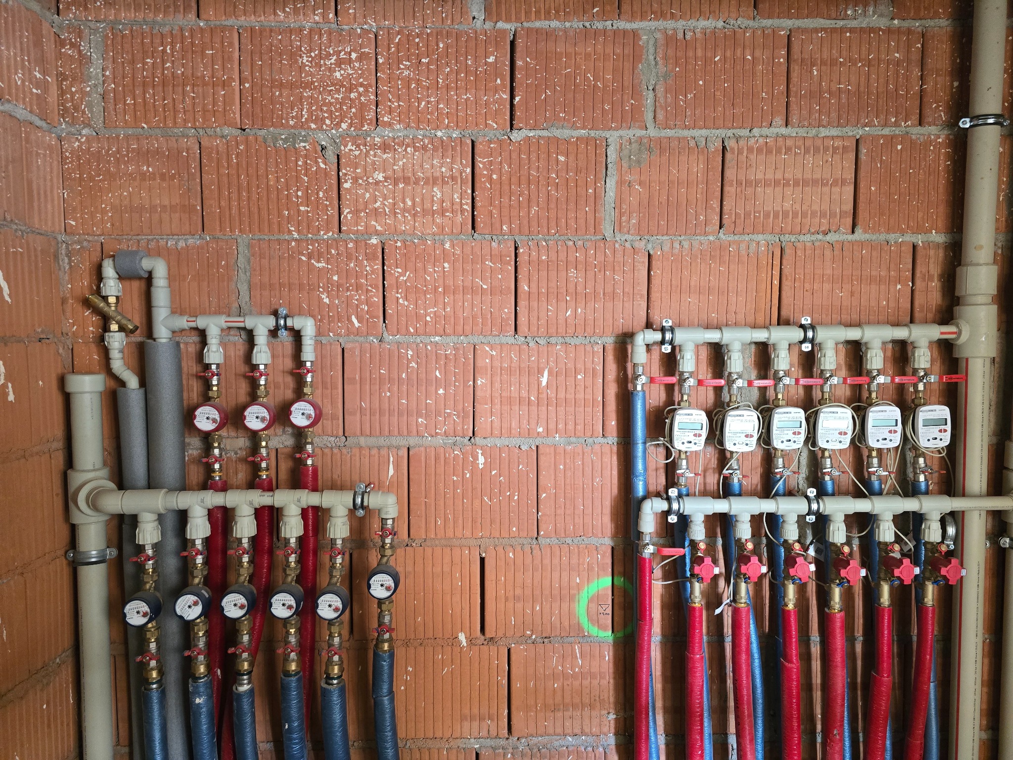 Rozdzielacz instalacji wodnej z licznikami na tle ściany z cegły dziurawki, widoczne rury w izolacji czerwonej i niebieskiej, oraz zawory.
