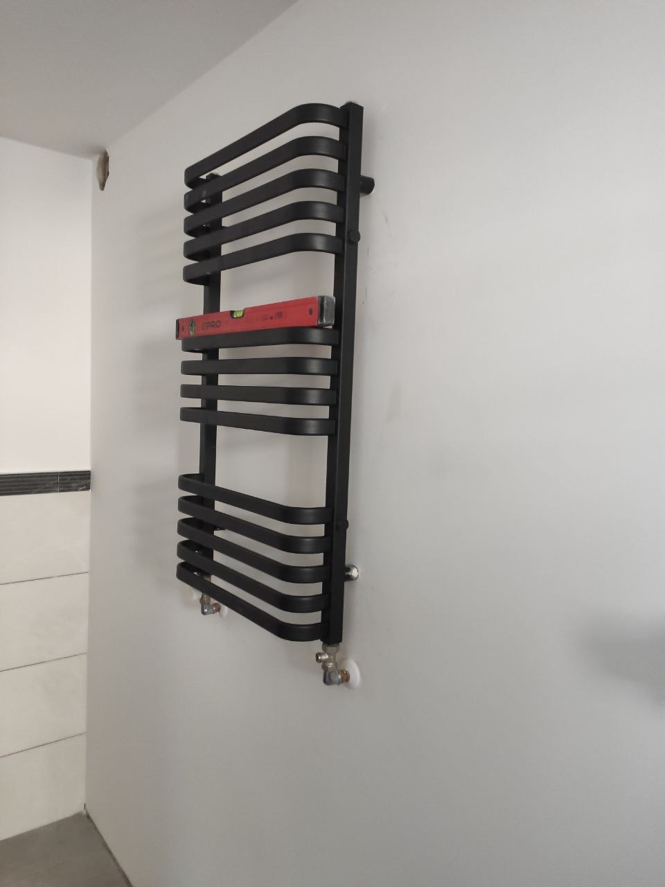 Czarny, designerski grzejnik łazienkowy drabinkowy, zamontowany na białej ścianie, poziomowany czerwoną poziomicą, widoczne przyłącza instalacji.