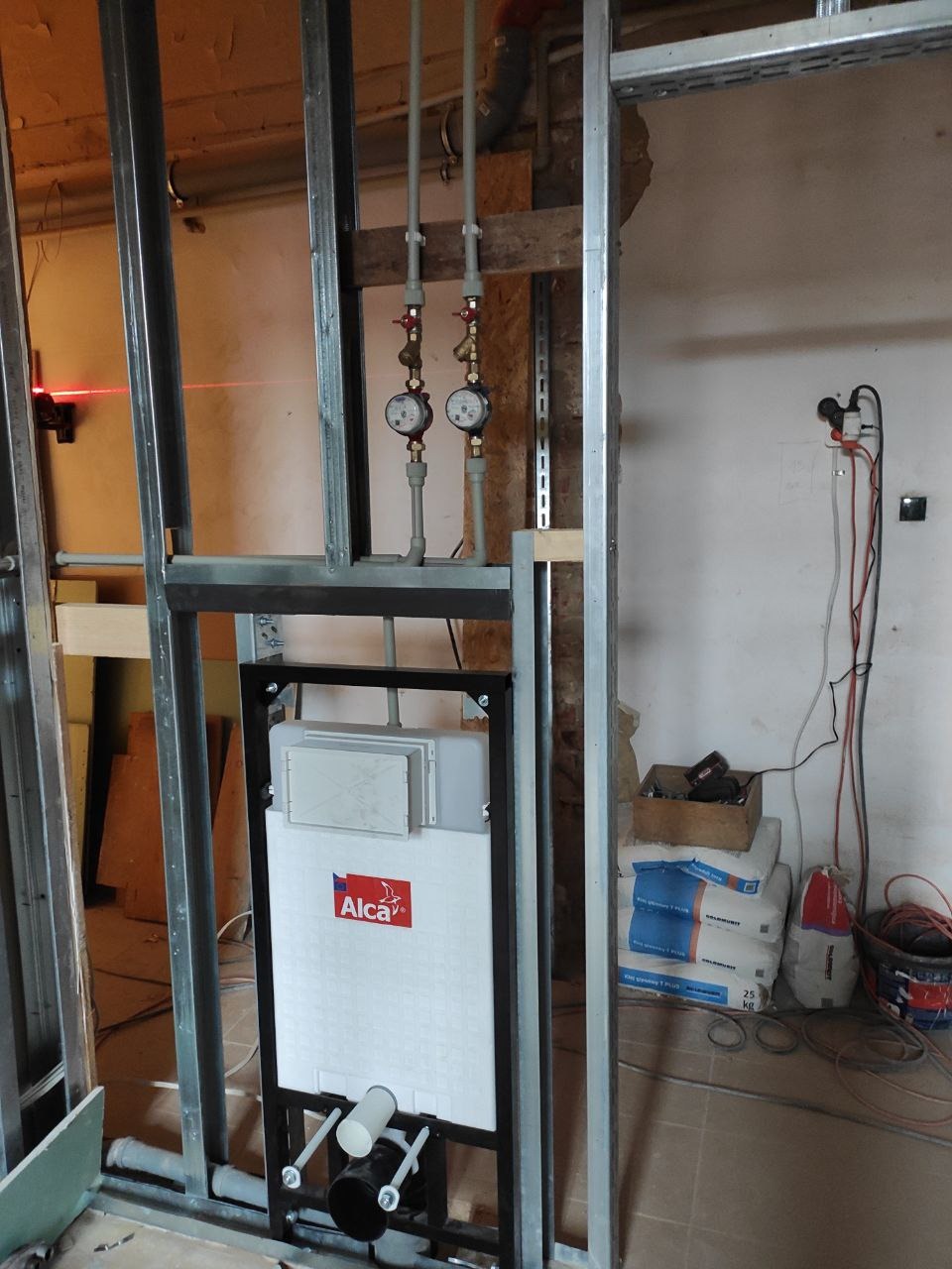 Stelaż podtynkowy WC marki Alca zamontowany w metalowej konstrukcji szkieletowej, widoczne rury doprowadzające wodę z zaworami i licznikami, laserowy niwelator na ścianie, worki z zaprawą murarską...