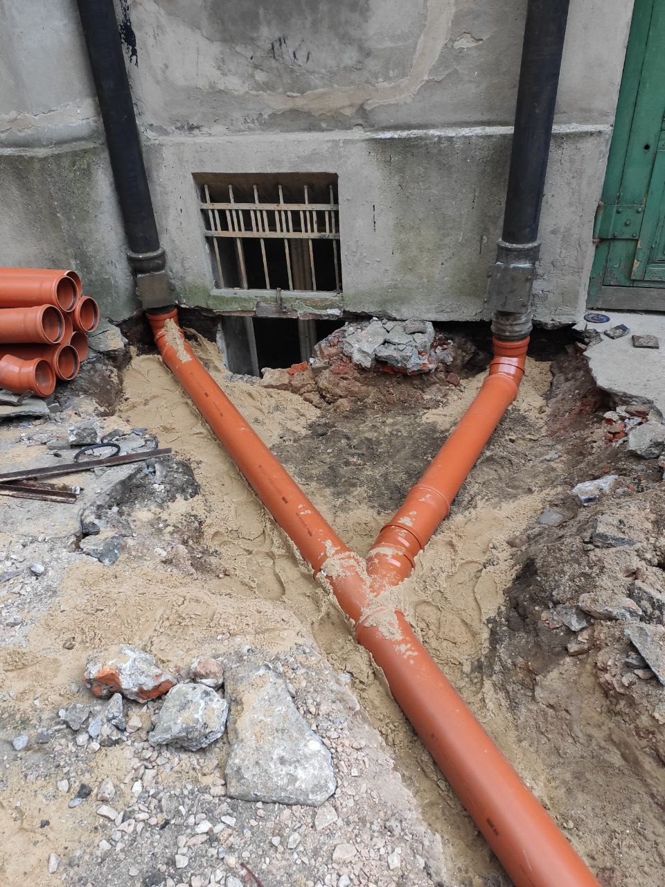 Wykonane przyłącze kanalizacyjne z rur PVC w wykopie, widoczne wejście do budynku z oknem zabezpieczonym kratą i fragmentem zielonych drzwi.