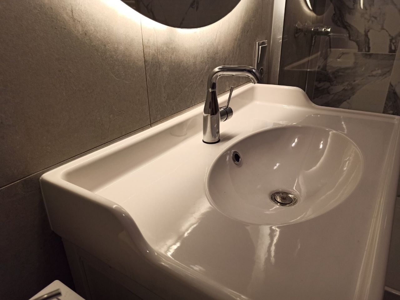 Biała umywalka nablatowa z chromowaną baterią, podświetlane lustro w tle, szare płytki na ścianie, minimalistyczny design.