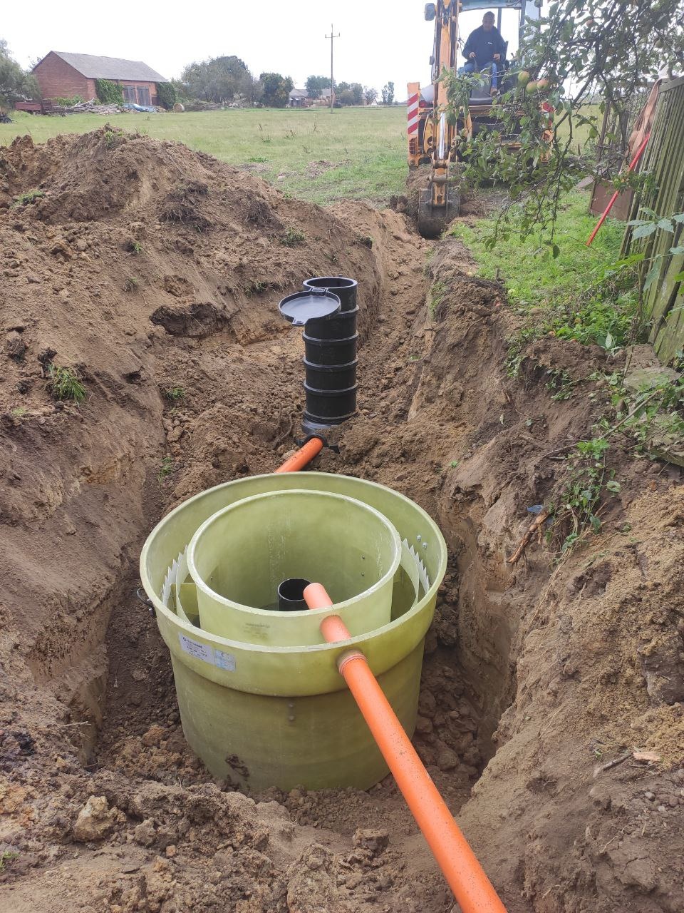 Instalacja przydomowej oczyszczalni ścieków w wykopie, widoczny zbiornik z zielonego tworzywa, czarna rura pionowa oraz pomarańczowa rura łącząca, w tle koparka i budynek mieszkalny.