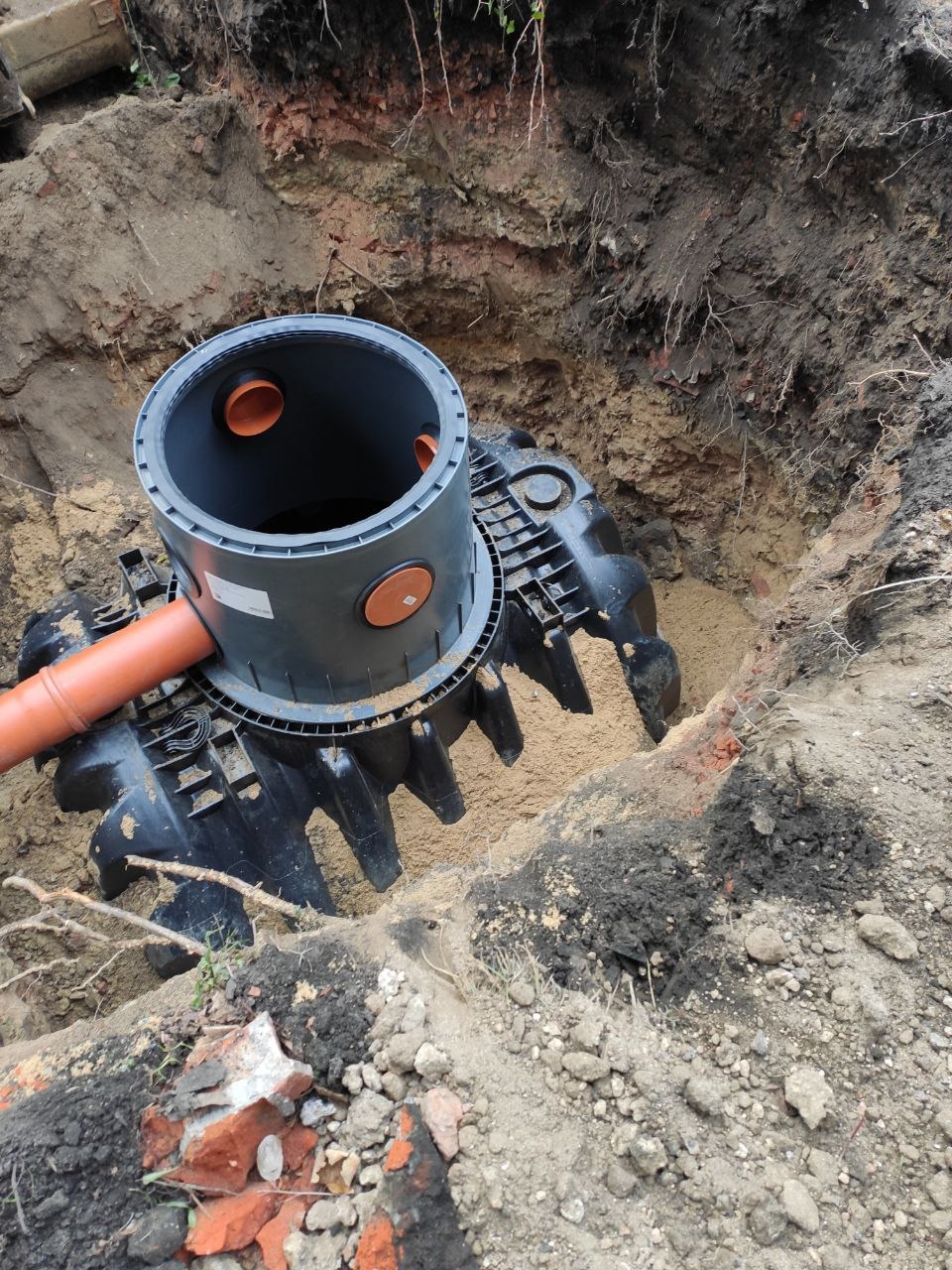 Szara studzienka kanalizacyjna z pomarańczowymi zaślepkami i rurą w wykopie ziemnym, widoczne korzenie i fragmenty cegieł.