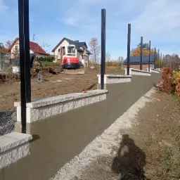 Wznoszona konstrukcja z betonu i kamienia z elementami stalowymi, prawdopodobnie w trakcie budowy, z widocznym mini-koparką Kubota w tle i domami jednorodzinnymi, cień fotografa w prawym dolnym rogu.