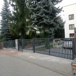 Nowoczesne, grafitowe ogrodzenie panelowe z bramą wjazdową i furtką, osadzone na niskim murku z szarej kostki, na tle zielonych drzew i białego budynku mieszkalnego.