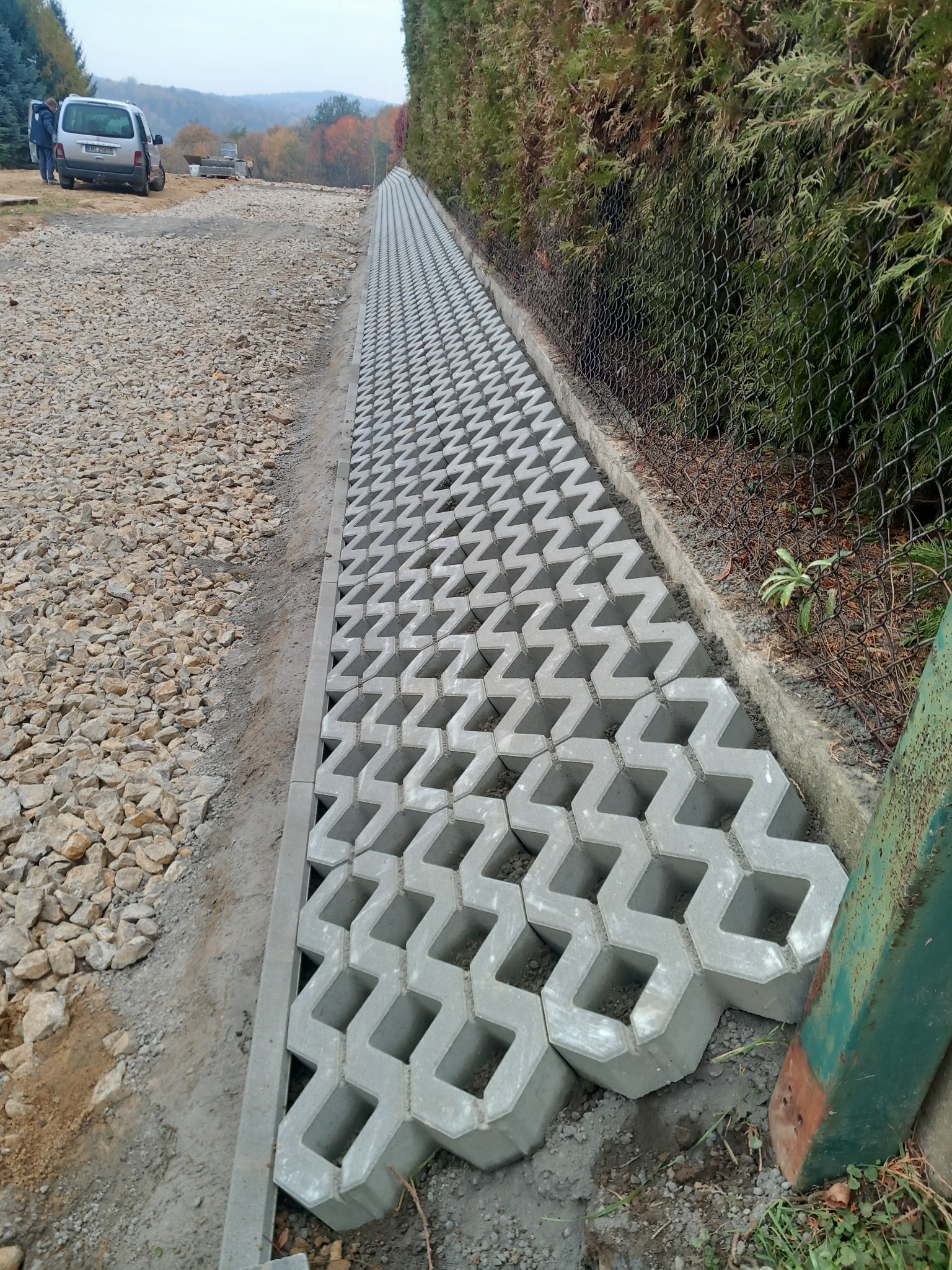 Ułożona ażurowa nawierzchnia z betonowych płyt w kształcie zygzaków, widok z perspektywy, obok wysypany kamień i siatka ogrodzeniowa obsadzona tujami.