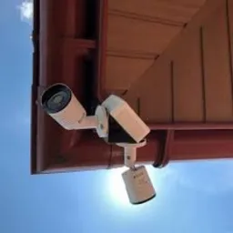 Dwie kamery monitoringu zamontowane pod okapem dachu w kolorze brązowym, widok z dołu na tle błękitnego nieba.