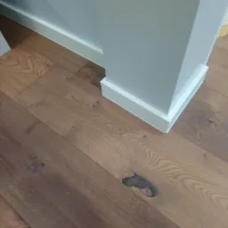 Fragment podłogi z ciemnego drewna z widocznymi słojami i sękiem, przy ścianie w jasnym odcieniu szarości z białą listwą przypodłogową i narożnikiem.