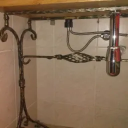 Kuta, metalowa konstrukcja pod umywalką z drewnianym blatem, widoczne elementy instalacji hydraulicznej i płytki ceramiczne w tle.