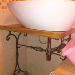 Jan Kohnke - Biała umywalka ceramiczna o owalnym kształcie, osadzona na drewnianym blacie wspartym na metalowej, dekoracyjnej konstrukcji w stylu retro, z widoczną armaturą i fragmentem mozaiki ściennej w tle.