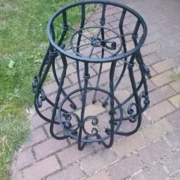 Kuty, czarny stojak na donicę z dekoracyjnymi zawijasami, umieszczony na kostce brukowej obok trawnika.