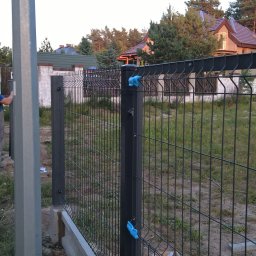 Jan Kohnke - Fragment montowanego ogrodzenia metalowego z podmurówką betonową, widoczne narzędzia w kartonie, tło z zielenią i domami.