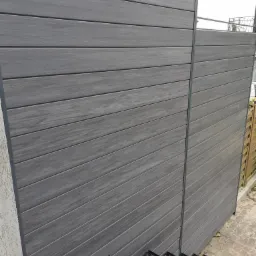 Szary panelowy system ogrodzeniowy z kompozytu drewna, widok z bliska na teksturę desek i metalowe słupki, częściowo widoczny taras kompozytowy na pierwszym planie.