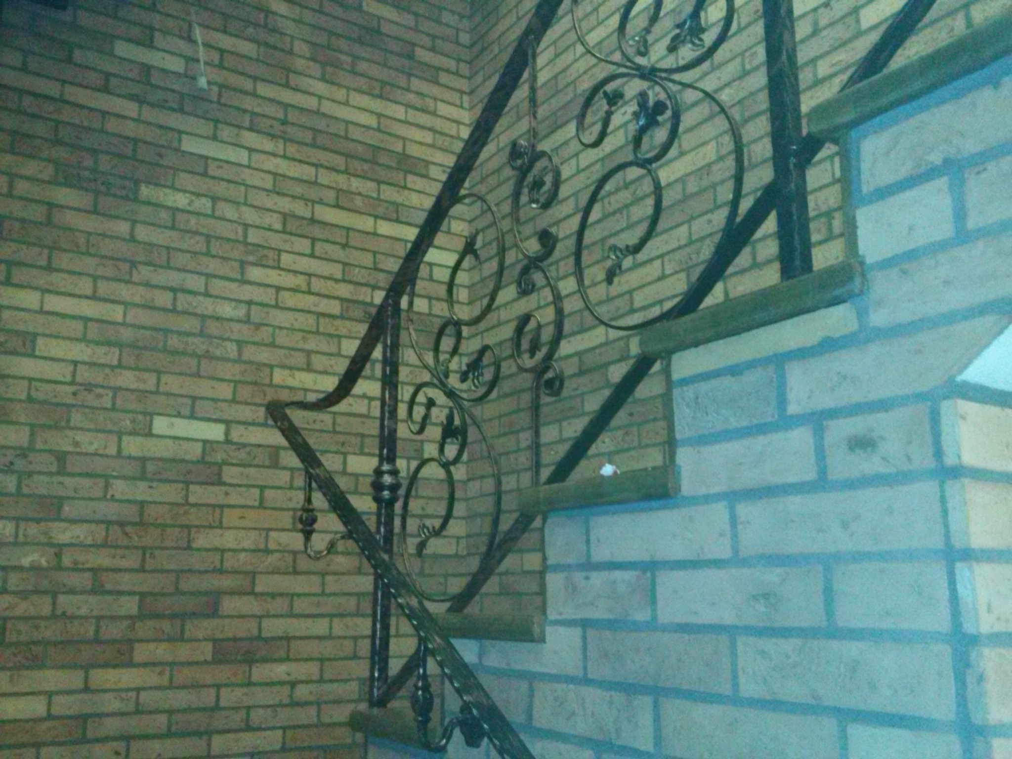 Kute, zdobione metalowe balustrady schodowe zamontowane przy ceglanej ścianie. Widoczne drewniane stopnie schodów.