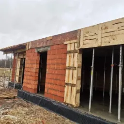 Wznoszenie ścian budynku z czerwonej cegły, widoczne szalunki drewniane i metalowe podpory, teren budowy w trakcie prac.