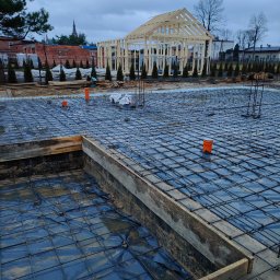 Usługi budowlane Szymon Gabryś - Zbrojenie fundamentów z widocznym szalunkiem i izolacją przeciwwilgociową, w tle konstrukcja szkieletowa domu w trakcie budowy, pochmurne niebo.