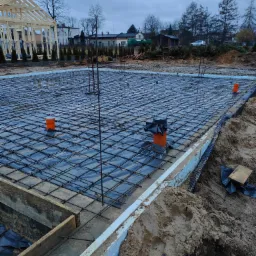 Przygotowanie zbrojenia płyty fundamentowej z widocznymi pionowymi prętami i pomarańczowymi rurami w Rybniku, na tle szkieletu drewnianego domu i pochmurnego nieba.