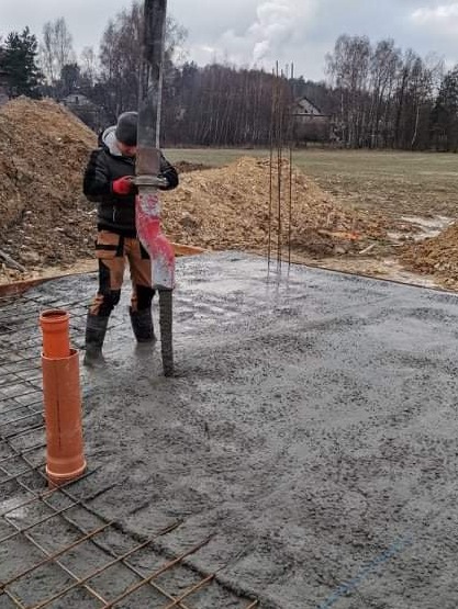 Pracownik w czerwonych rękawicach wylewa beton z pompy na przygotowane zbrojenie pod fundament, widoczny pomarańczowy pion kanalizacyjny, w tle hałda ziemi i drzewa.