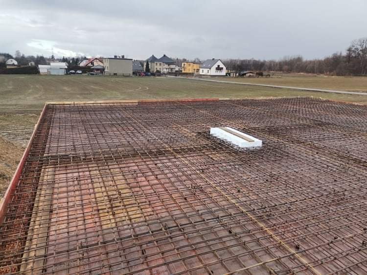Przygotowanie zbrojenia płyty fundamentowej z widoczną warstwą izolacji termicznej w centralnej części, na tle zabudowy jednorodzinnej i pochmurnego nieba.