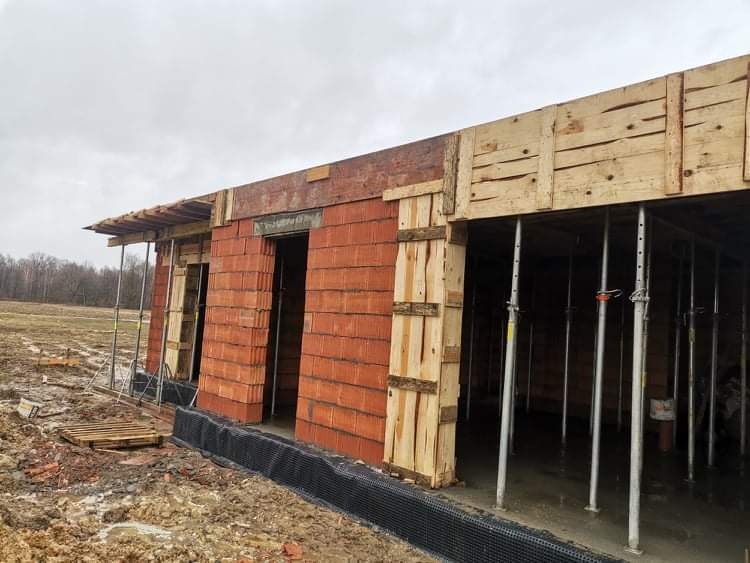 Wznoszenie ścian budynku z czerwonej cegły, widoczne szalunki drewniane i metalowe podpory, teren budowy w trakcie prac.