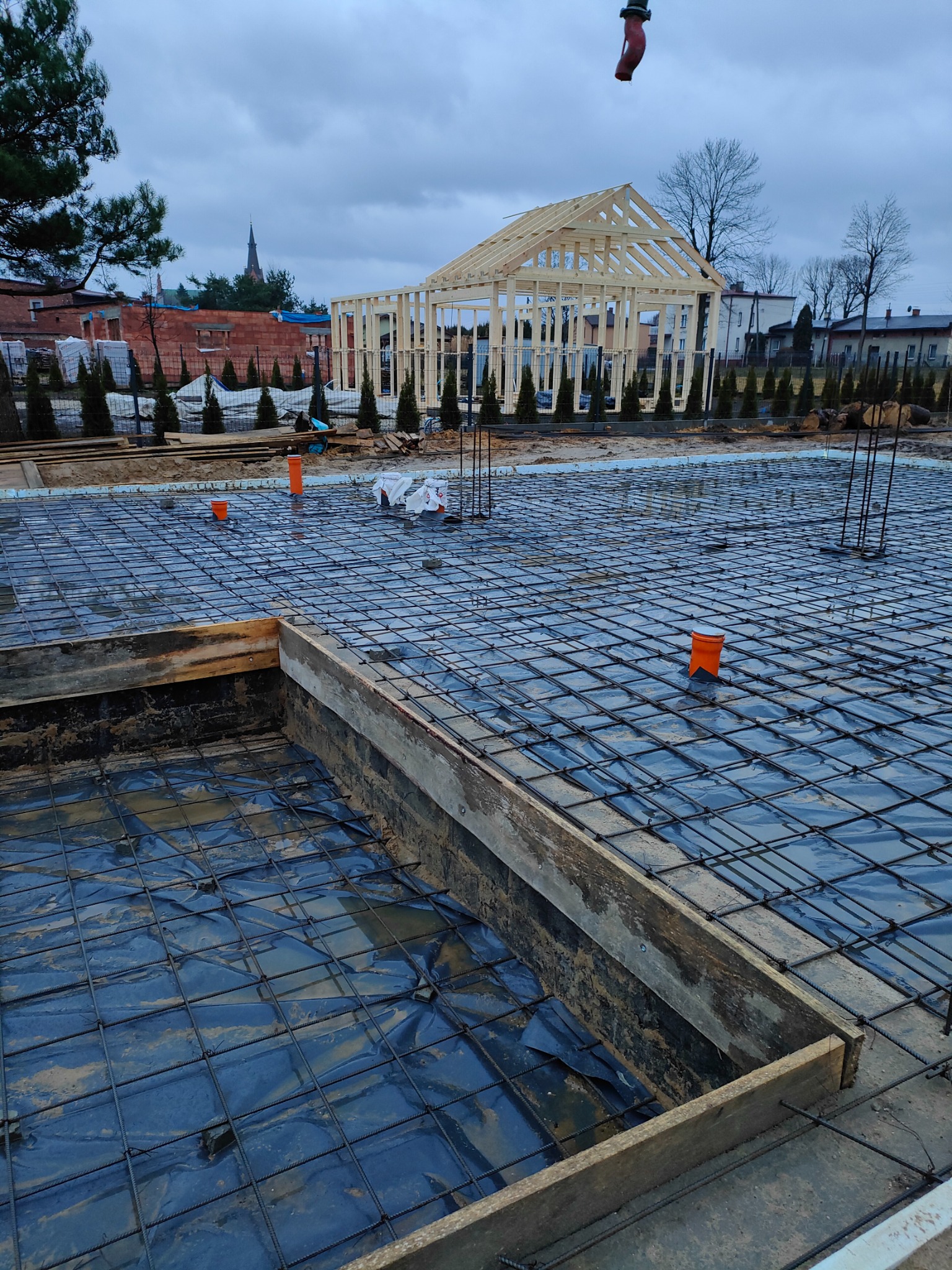 Zbrojenie fundamentów z widocznym szalunkiem i izolacją przeciwwilgociową, w tle konstrukcja szkieletowa domu w trakcie budowy, pochmurne niebo.
