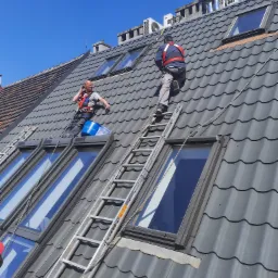 Dwóch monterów na dachu z szarej blachodachówki trapezowej, jeden rozmawia przez telefon, drugi wspina się po drabinie w kierunku komina i klimatyzatorów, obaj zabezpieczeni uprzężą.