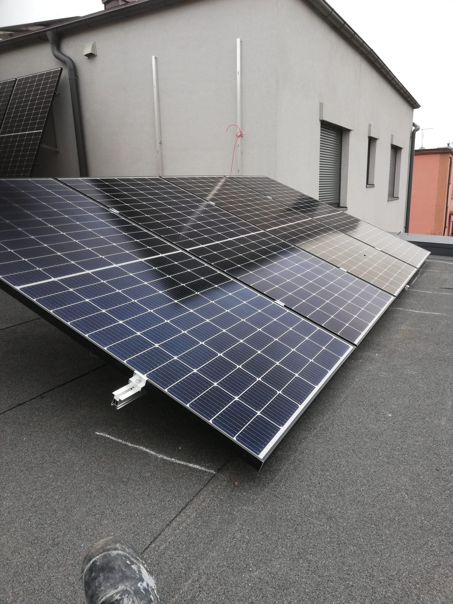 Panele słoneczne zamontowane na płaskim dachu budynku, widoczne elementy mocujące i fragment buta pracownika w dolnym rogu.