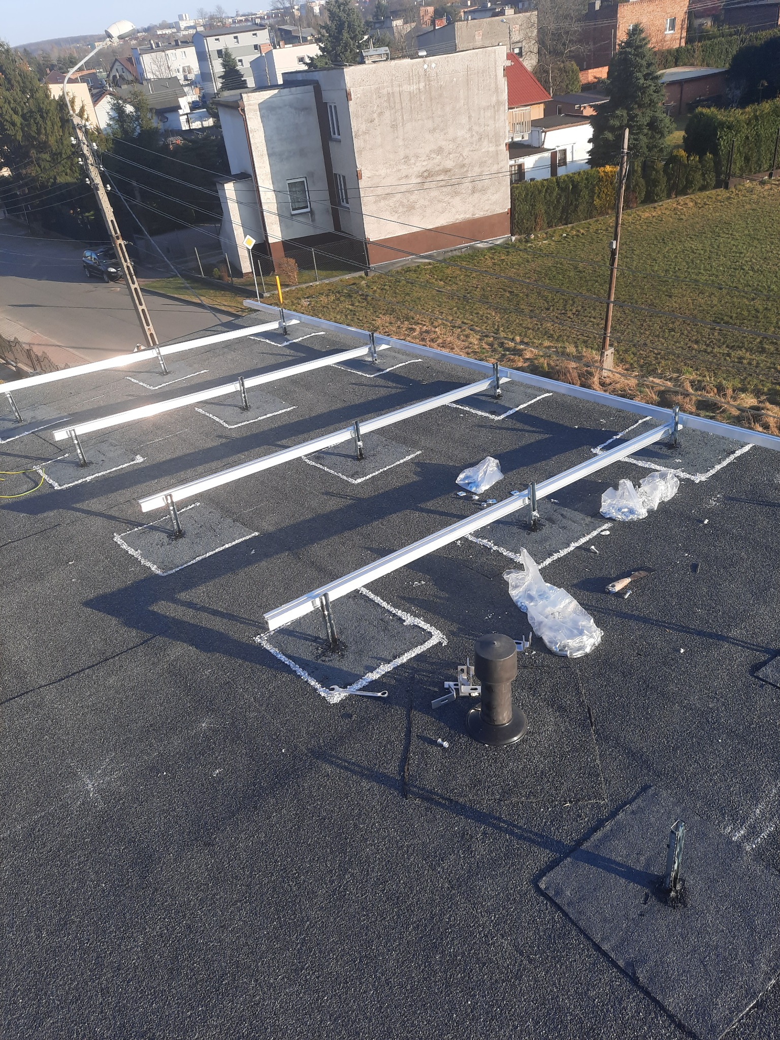 Montaż konstrukcji wsporczej pod panele słoneczne na płaskim dachu pokrytym papą, widoczne elementy mocujące i narzędzia.