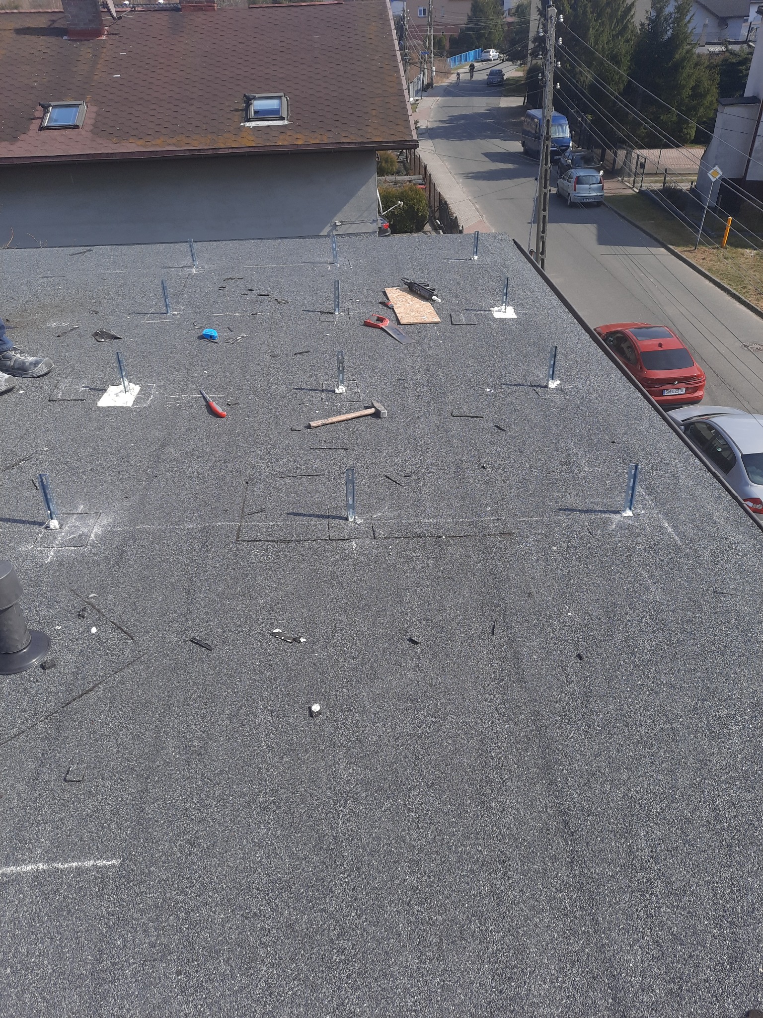 Montaż konstrukcji wsporczej na dachu pokrytym papą, przygotowanie do instalacji paneli słonecznych, widoczne narzędzia i elementy montażowe, perspektywa z góry.