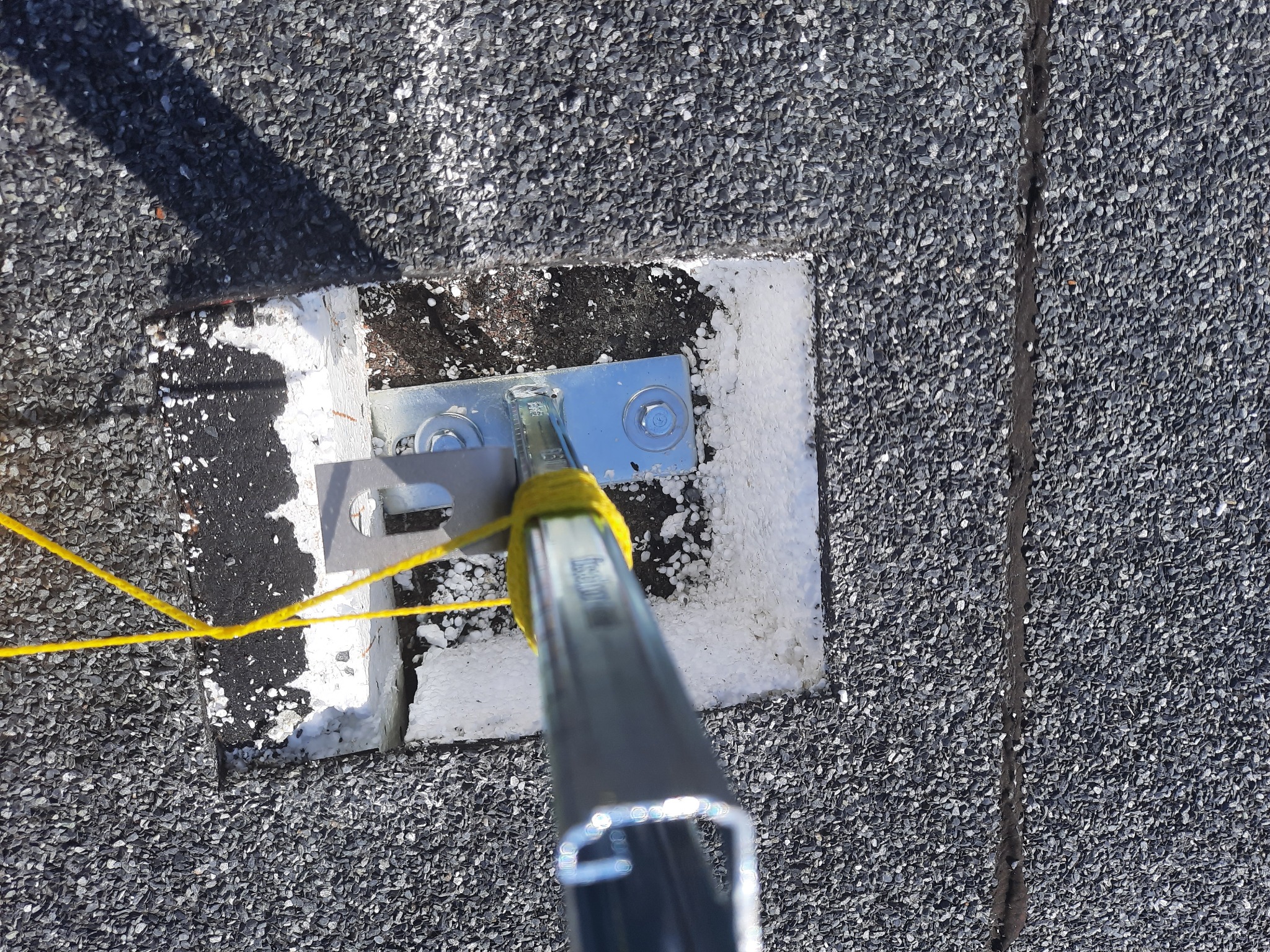 Mocowanie konstrukcji wsporczej na dachu pokrytym gontem bitumicznym, zabezpieczone żółtą taśmą. Widoczna izolacja wokół elementu mocującego.