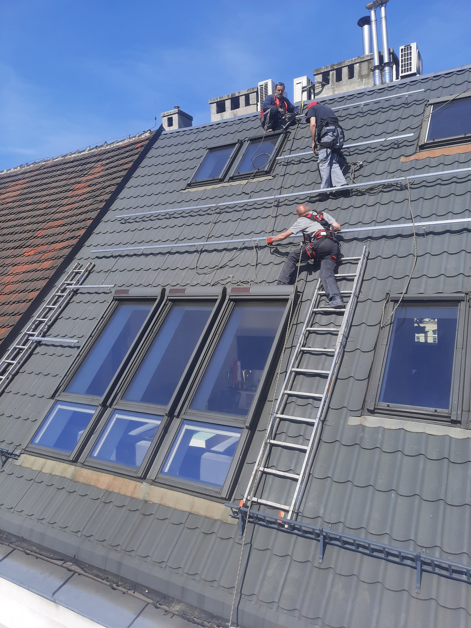 Montaż konstrukcji pod panele na dachu z oknami dachowymi, pracownicy zabezpieczeni linami i uprzężami, jeden z nich wchodzi po drabinie.