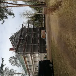 Dom jednorodzinny w trakcie ocieplania elewacji, widoczne rusztowania, ocieplona szara ściana, pracownik na rusztowaniu, drzewa w otoczeniu, pochmurne niebo.