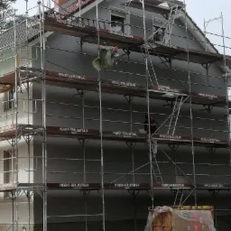 Dom jednorodzinny w trakcie tynkowania elewacji, widoczne rusztowania z logo Menke-Geruestbau, pracownik na drabinie i pojemniki z zaprawą tynkarską