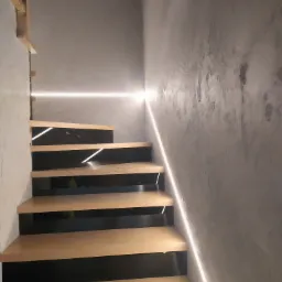 Nowoczesne schody z podświetleniem LED, drewniane stopnie z czarnymi podstopnicami, ściana wykończona dekoracyjnym tynkiem.