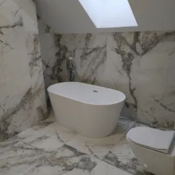 Nowoczesna łazienka na poddaszu z marmurowymi płytkami, wolnostojącą białą wanną i toaletą podwieszaną, oświetlona przez okno dachowe.