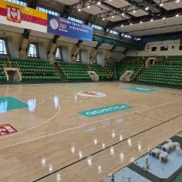 Sala gimnastyczna z parkietem po cyklinowaniu, widoczne odświeżone logo i linie boiska. Na podłodze błyszczy świeży lakier. Trybuny z zielonymi krzesełkami.
