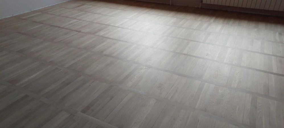 Jasny, odnowiony parkiet ułożony w kwadraty, widoczne ślady po szlifowaniu i jasne fugi.
