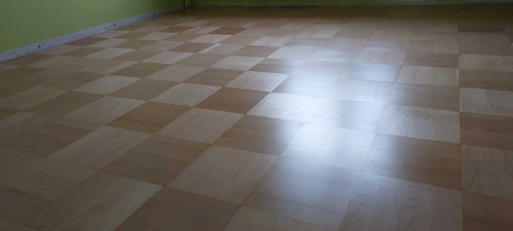 Ułożona w szachownicę podłoga z parkietu w odcieniach jasnego drewna, widoczne odbicia światła na powierzchni.