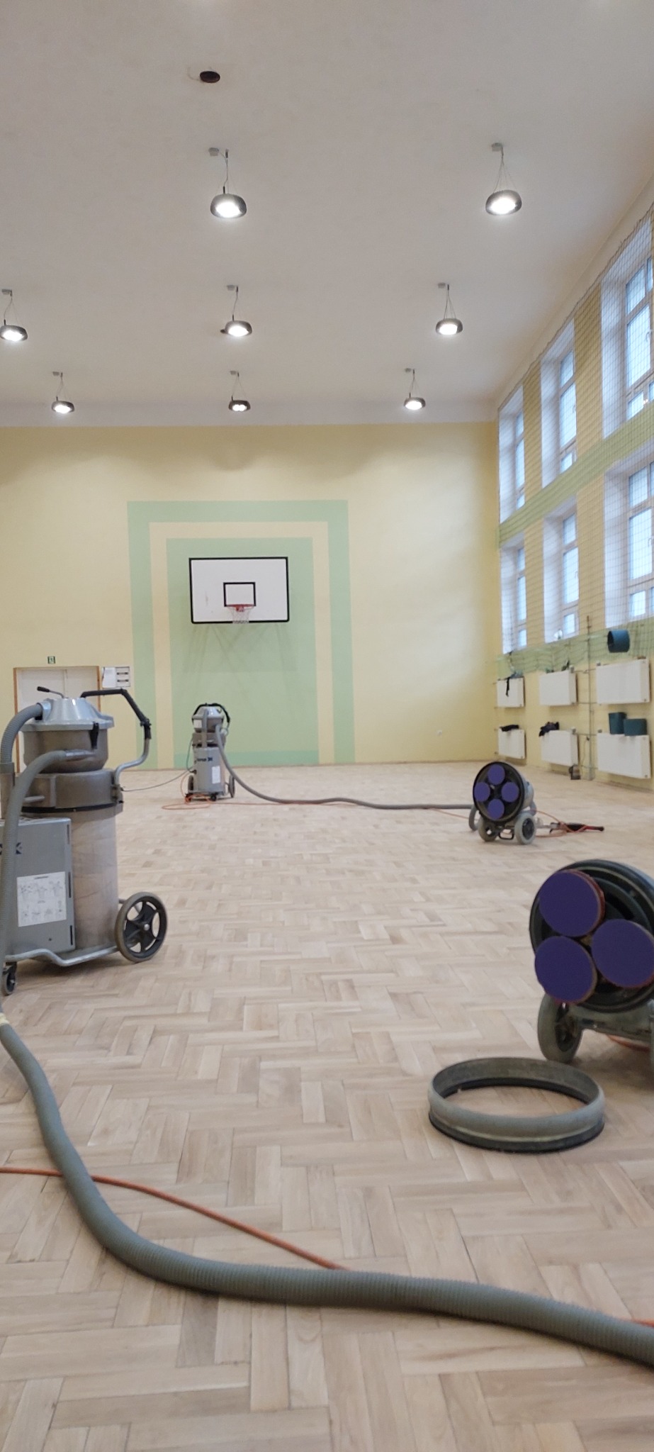 Sala gimnastyczna z parkietem w jodełkę podczas prac renowacyjnych, widoczne maszyny do szlifowania oraz odkurzacz przemysłowy, na ścianie kosz do koszykówki.