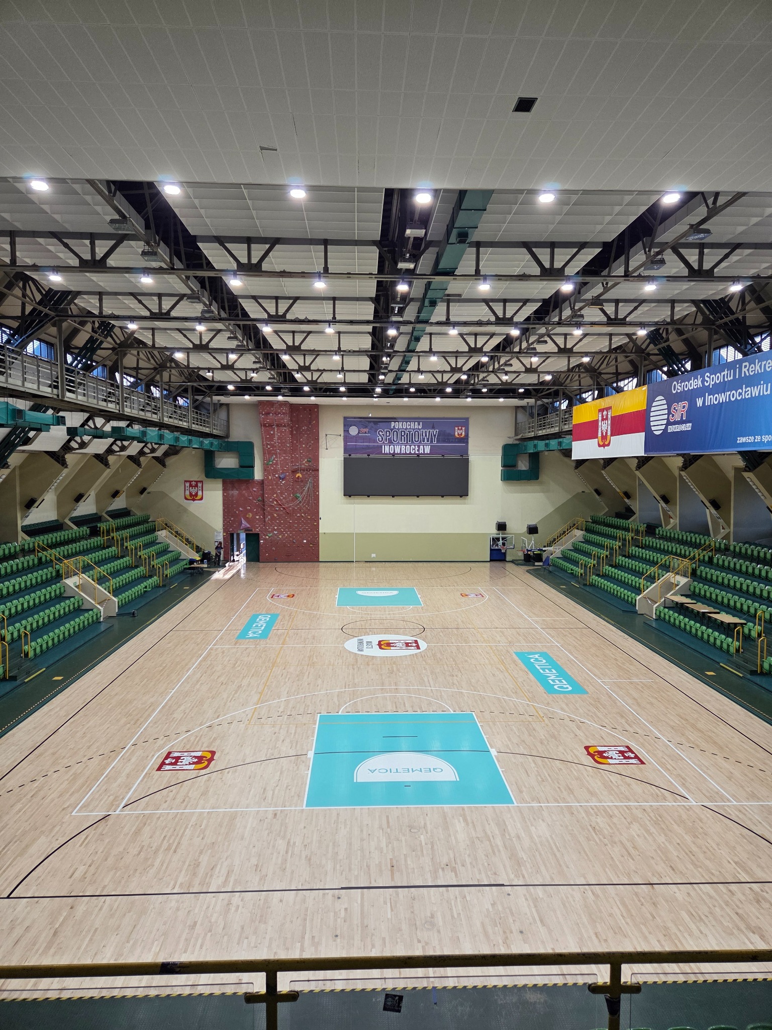 Widok z góry na parkiet hali sportowej z zielonymi trybunami. Parkiet z jasnego drewna z oznaczeniami boiska i logo. W tle ściana wspinaczkowa i banery informacyjne.