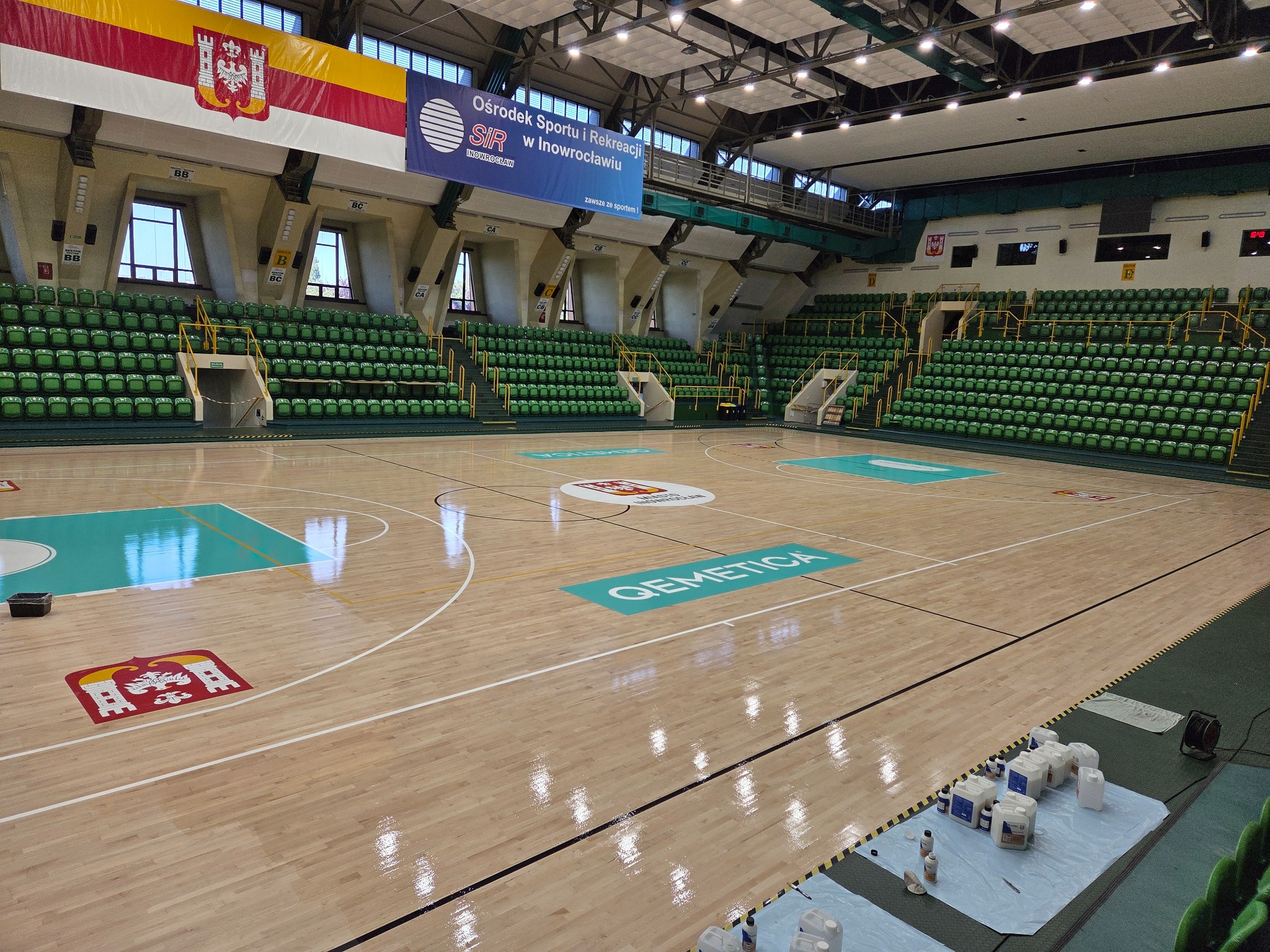 Sala gimnastyczna z parkietem po cyklinowaniu, widoczne odświeżone logo i linie boiska. Na podłodze błyszczy świeży lakier. Trybuny z zielonymi krzesełkami.