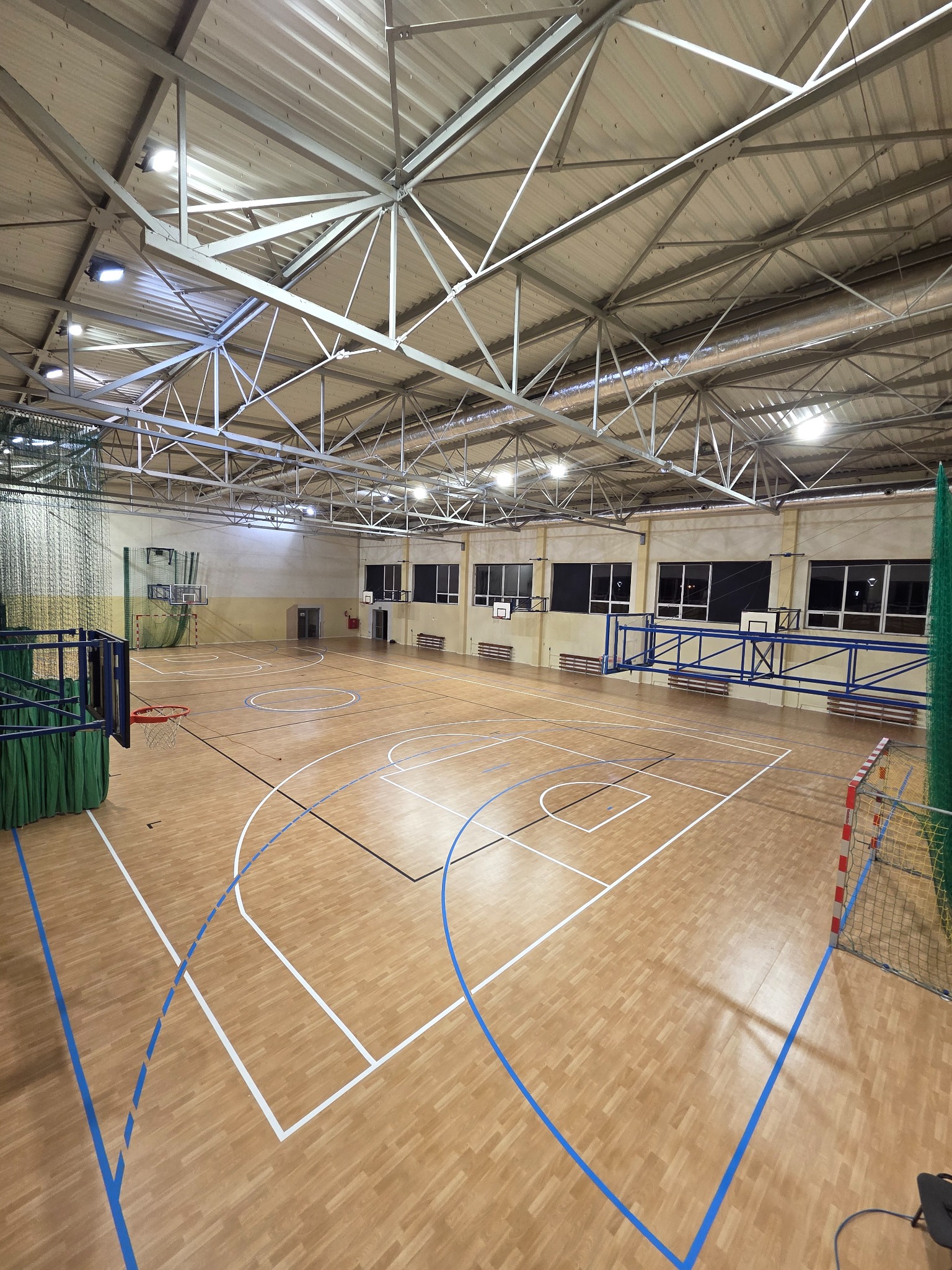 Sala gimnastyczna z parkietem w odcieniach beżu, z wyraźnie oznaczonymi liniami boiska w kolorze białym i niebieskim. Widoczne kosze i bramki do piłki ręcznej.