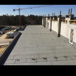 Widok z góry na płaski dach pokryty papą termozgrzewalną na nowo budowanym budynku, z widocznymi kominami wentylacyjnymi, obróbkami blacharskimi i żurawiem budowlanym w tle.