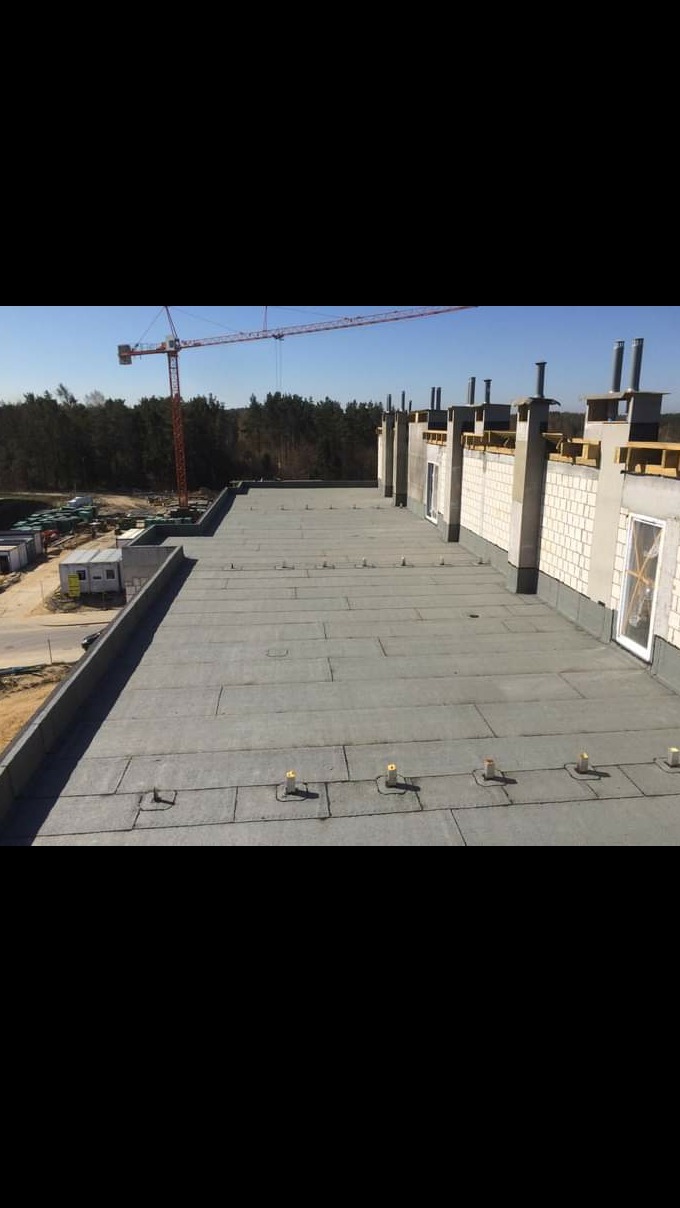 Widok z góry na płaski dach pokryty papą termozgrzewalną na nowo budowanym budynku, z widocznymi kominami wentylacyjnymi, obróbkami blacharskimi i żurawiem budowlanym w tle.