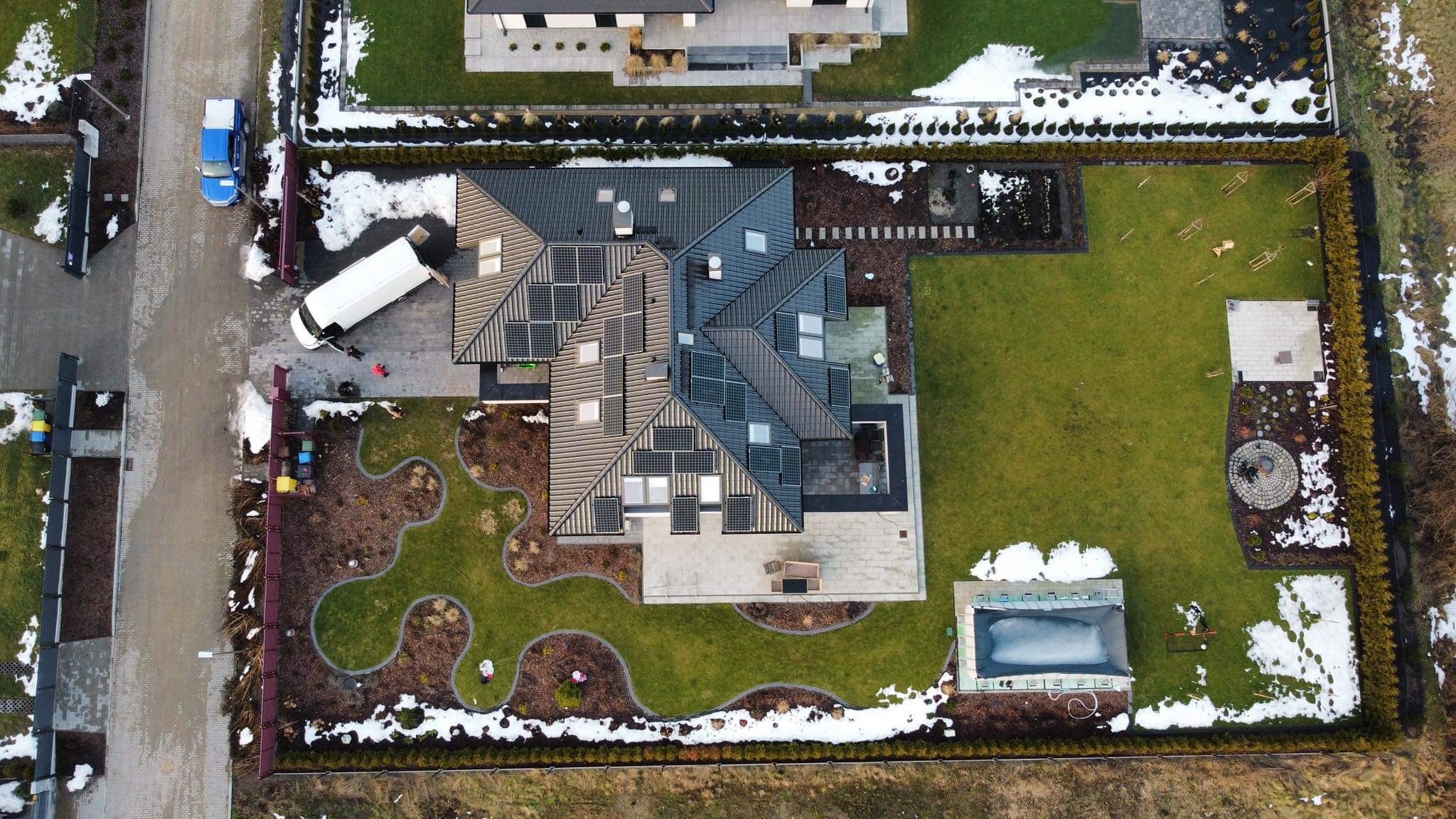 Widok z drona na dom jednorodzinny z panelami fotowoltaicznymi na dachu, częściowo pokrytym śniegiem, z białym busem dostawczym zaparkowanym obok, na tle zadbanego ogrodu z basenem i elementami...
