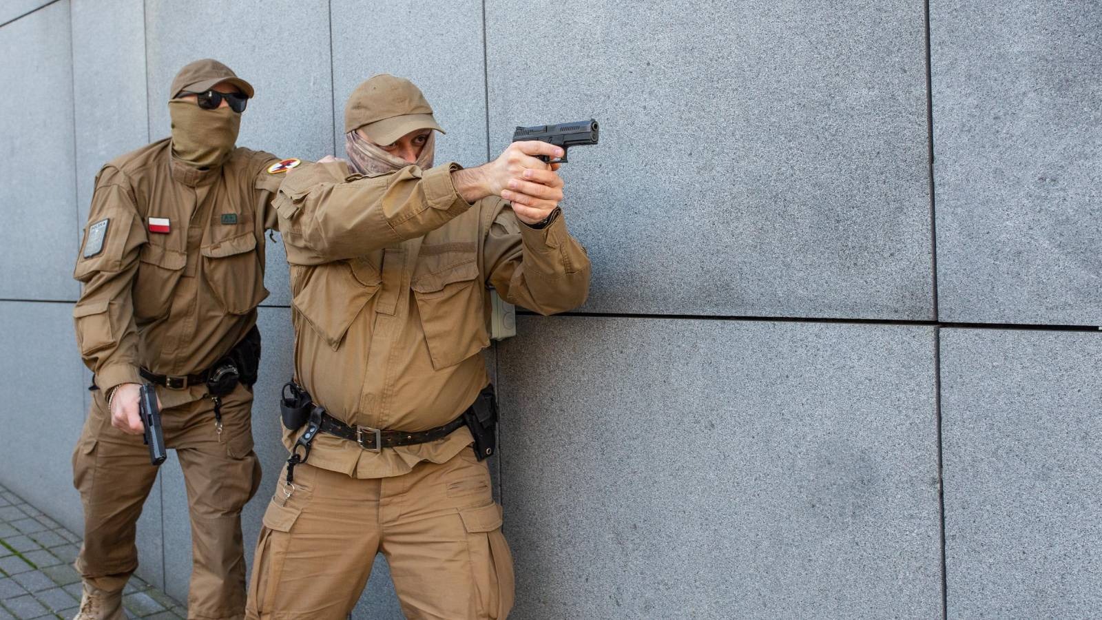 Dwóch uzbrojonych mężczyzn w kamuflażu, jeden celuje z pistoletu, obaj stoją przy szarej ścianie.
