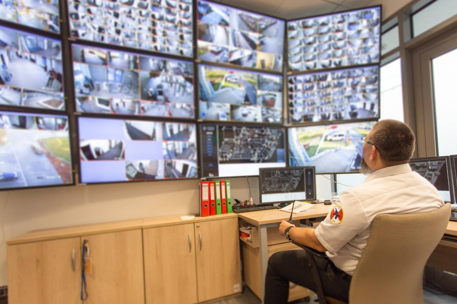Mężczyzna siedzi przed ścianą monitorów z podglądem z kamer CCTV, na blacie biurka radiotelefon i dokumenty.